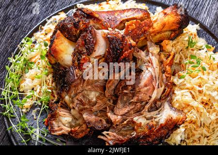 Knuspriger Schweinsknöchel, gebratener Schinkenbock, Schweinshaxe, mit Sauerkraut auf schwarzer Platte auf dunklem Holztisch, horizontale Ansicht von oben Stockfoto