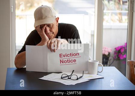 Trauriger Mann mit Mütze, der Evction Note anschaut, während er seinen Kopf in den Händen hält. Frustriert über Hypothekenzahlungen und Finanzverschuldung. Aus dem Hotel vertrieben werden Stockfoto