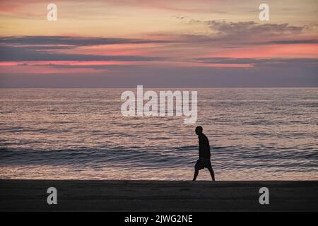 Ein einsamer Mann in Silhouette geht bei Sonnenaufgang in Einsamkeit am Strand entlang. Stockfoto