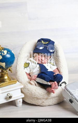 Kleinkind Junge im niedlichen Piloten Kostüm liegen auf weichen winzigen Stuhl kleine Kinder Requisiten Reise Koffer Globus Statue auf Tisch Stockfoto