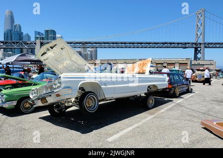 Ein weißer Lowrider-Wagen des Chevy Impala aus dem Jahr 1964 mit erhöhter Motorhaube und Hydrauliksystem, der im Hintergrund mit der Bay Bridge in San Francisco, Kalifornien, besetzt ist. Stockfoto