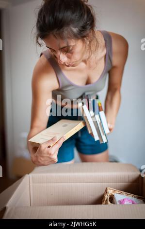 Eine entspannte junge Frau nimmt verschiedene Arten von Büchern aus einer Schachtel in ihrem Zimmer Stockfoto