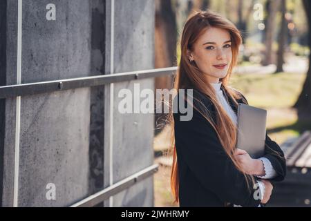 Schöne Geschäftsfrau, die im Stadtpark mit einem Laptop steht und auf ihre Freundin wartet, die zur Arbeit geht. Fröhliche Freiberuflerin. Büroarbeit Stockfoto