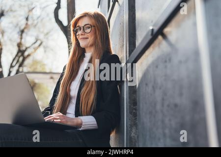 Schöne Geschäftsfrau, die auf der Bank sitzt und Remote-Arbeiten im Freien mit einem Laptop macht. Im Internet surfen und nach Informationen suchen. Freiberuflicher Mitarbeiter Stockfoto