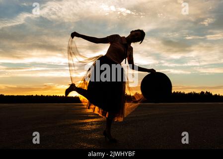 Tanzende Frau mit Hut in der Hand versteckt spielerisch die untergehende Sonne hinter dem Rock von Ballerina vor dem Hintergrund der Silhouette des Waldes und des wolkigen Sonnenuntergangs Stockfoto