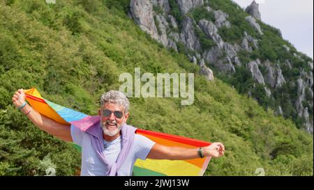 Porträt eines grauhaarigen älteren kaukasischen Mannes Bisexualität mit Bart und Sonnenbrille mit einer Regenbogenflagge über der Natur. Feiert den Pride-Monat, Rainbow Flag Day, Gay Parade Stockfoto