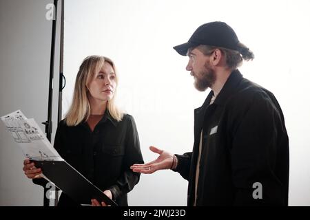 Junge blonde Assistentin des Fotografen mit Dokumenten, die Details von Video- oder Fotoaufnahmen mit ihm im Studio besprechen Stockfoto