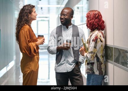 Drei junge erfolgreiche Büromanager in Casualwear, die sich zwischen zwei Wänden im Korridor eines modernen Gebäudes unterhalten Stockfoto