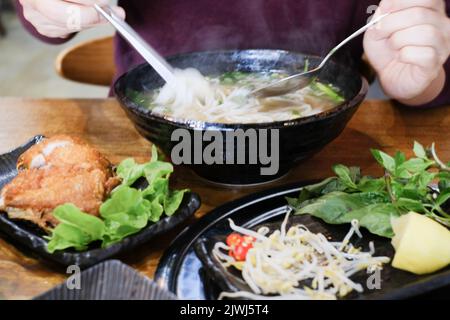 Mann, der eine Schüssel mit Phở Bò (Rindfleisch-Pho) und Gà Da Dòn (knusprig-hautiges Huhn) im Me Pho, einem vietnamesischen Restaurant in Bankstown – Sydney, Australien, isst Stockfoto