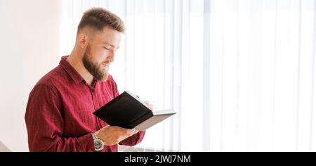 Der bärtige Mann aus dem Geschäft steht gegen ein Fenster im Büro und schreibt Pläne in einem Notizbuch in einem dunklen burgunderfarbenen Hemd auf. Konzept-Banner Stockfoto