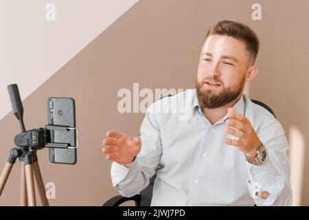 Lächelnder bärtiger Geschäftsmann, der seine Video-Blog-Episode mit dem Smartphone filmt, am Schreibtisch sitzt, schreibt in ein Notebook und gestikuliert mit seiner Hand. In einem Hemd gekleidet Stockfoto