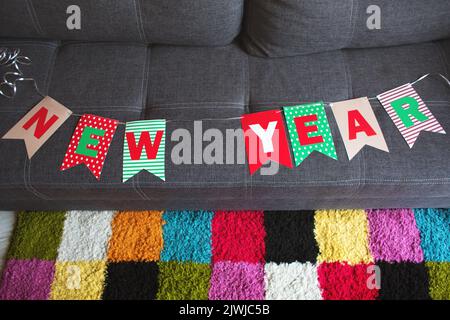 Eine Girlande aus Fahnen mit der Aufschrift Happy New Year, auf einem grauen Sofa vor dem Hintergrund eines farbigen Teppichs Stockfoto