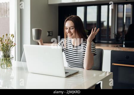 Junge lachende und fröhliche Frau mit weißem Laptop und Kaffee trinken. Videoanruf, Webinar, Konferenz, Remote-Job Stockfoto