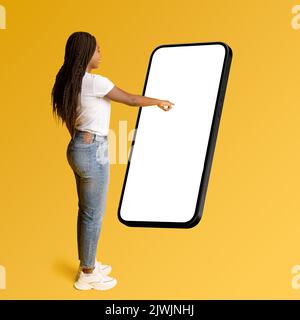 Neue Werbung für mobile Apps. Die afroamerikanische Frau berührt den riesigen Smartphone-Bildschirm, wählt etwas aus, scrollt auf dem großen leeren Display des Mobiltelefons, kopiert Speicherplatz, macht Mockup Stockfoto