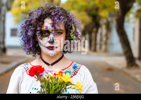 Vorderansicht Porträt einer ernsthaften jungen Frau mit Lockenwicklern, die Blumen halten und mit künstlerischem Make-up, das La Calavera Catrina darstellt, im Freien stehend l Stockfoto