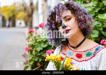 lateinamerikanische argentinische junge Frau mit La Calavera Catrina Make-up und folklorischer Kleidung, die mit einem nachdenklichen Gesicht auf die Kamera schaut und Blumen hält, ist s Stockfoto