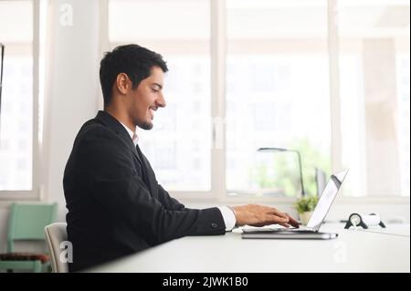 Optimistischer hispanischer Büroangestellter mit Laptop im modernen Coworking Space, Geschäftsmann in formellem Anzug, der am Computer arbeitet, am Bildschirm zuschaut und lächelt, Seitenansicht Stockfoto