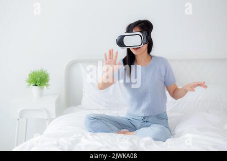 Junge asiatische Frau mit vr-Headset spielen Spiel mit aufgeregt auf dem Bett im Schlafzimmer zu Hause, weiblich mit Virtual Reality oder Metaverse Innovation für Stockfoto