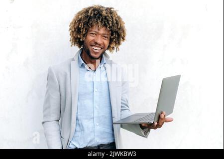 Gutaussehender positiver erfolgreicher, lockiger Latino oder brasilianischer Mann, stilvoll elegant gekleidet, mit offenem Laptop in der Hand, während er auf isoliertem Hintergrund steht, die Kamera anschaut und freundlich lächelt Stockfoto