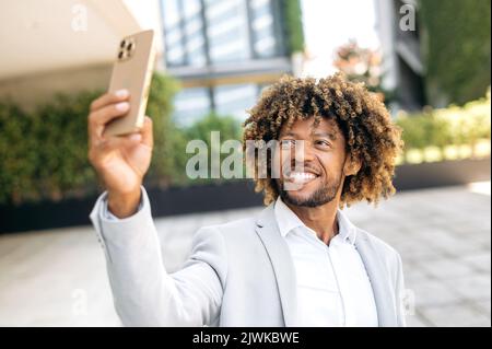 Selfie-Foto-Konzept. Positiver gutaussehender, lockiger, gemischter Rennmensch, der im Freien steht, ein Selfie auf ihrem Smartphone macht, ein gemischter Blogger, der Live-Streaming im sozialen Netzwerk hat und lächelt Stockfoto