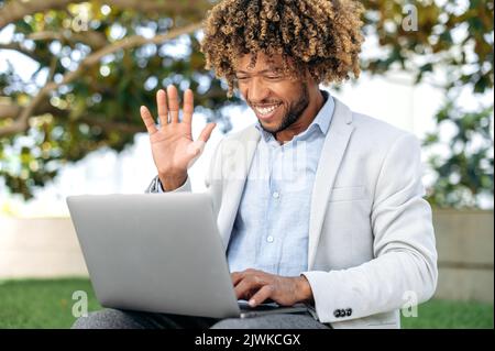 Online-Videokommunikation. Positive gemischte Rasse lockiger Mann, Mitarbeiter des Unternehmens, Mentor, mit Laptop für Videokonferenz-Gespräch mit Kollegen oder Kunden, Begrüßung Gesprächspartner, lächelnd Stockfoto