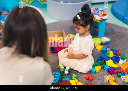 Spielerisch im Kindergarten lernen. Kleinkind kleines Mädchen und die Lehrerin spielen mit bunten Kunststoff-Spielhäusern, Bausteinen, Autos und Booten. Entwicklung von Phantasie, Kreativität, Feinmotorik und grobmotorischen Fähigkeiten. Hochwertige Fotos Stockfoto