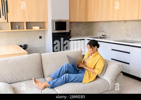Entspannte junge afroamerikanische Freiberuflerin sitzt auf der Couch und schaut sich Filmserien auf dem Laptop an oder arbeitet aus der Ferne, tippt E-Mails, Seitenansicht Stockfoto