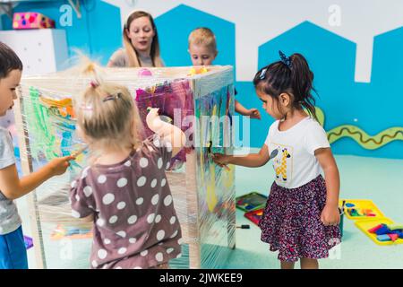 Frischhaltefolie Malerei für die Verbesserung der Kinder Phantasie und Gehirnentwicklung. Kleinkinder und ihre Lehrerin malen mit Bürsten, Rollen und Farben auf einer Frischhaltefolie, die um den hölzernen Regalständer gewickelt ist. Lustige Aktivität für Kinder feine und grobe Motorik im Kindergarten. Hochwertige Fotos Stockfoto