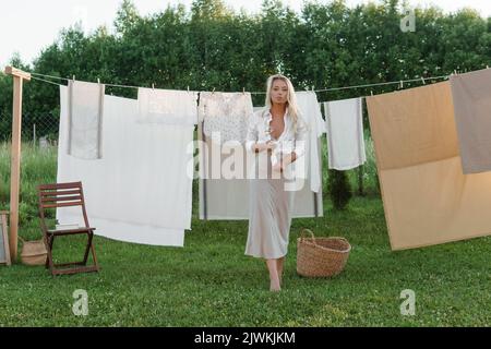 Wäschereitag. Eine Frau hängt Bettwäsche und Handtücher auf einem Baum im Hof eines Dorfhauses. Sommerhaus-Konzept. Stockfoto