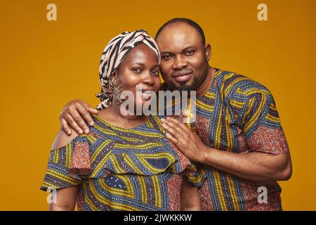 Porträt eines hübschen afrikanischen Paares in Nationalkostümen, umarmt und lächelnd auf die Kamera vor gelbem Hintergrund stehend Stockfoto