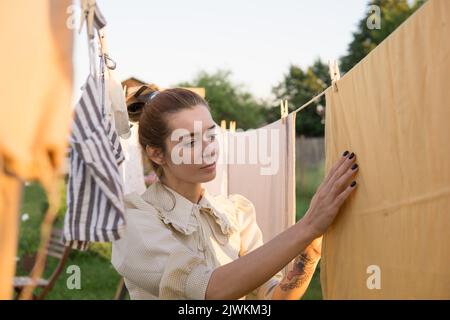 Wäschereitag. Eine Frau hängt Bettwäsche und Handtücher auf einem Baum im Hof eines Dorfhauses. Sommerhaus-Konzept. Stockfoto