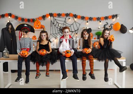 Kinder, die in verschiedenen gruseligen Figuren gekleidet sind, sitzen in einer Reihe und halten Kürbiseimer für Süßigkeiten. Stockfoto