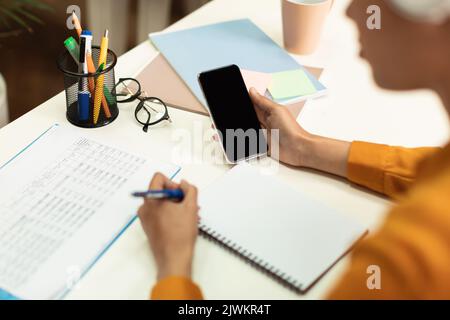 Lateinische Studentin hält Smartphone mit leerem Bildschirm und schreibt Notizen im Notizbuch, studiert online in der Bibliothek Stockfoto