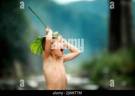 Der Junge unter der Klette. Kind versteckt sich vor dem Regen unter den Blättern und lacht. Spiele im Freien Stockfoto