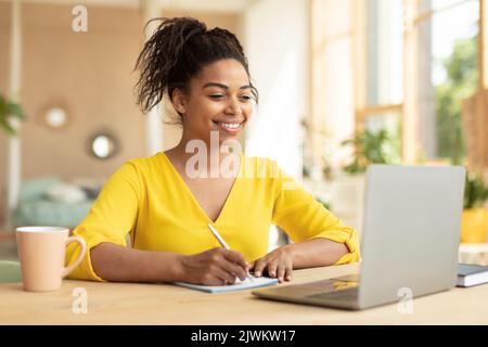 Lächelnde afroamerikanische Dame, die am Laptop arbeitet, Notizen im Notebook macht, zu Hause am Schreibtisch sitzt und Platz für Kopien hat Stockfoto