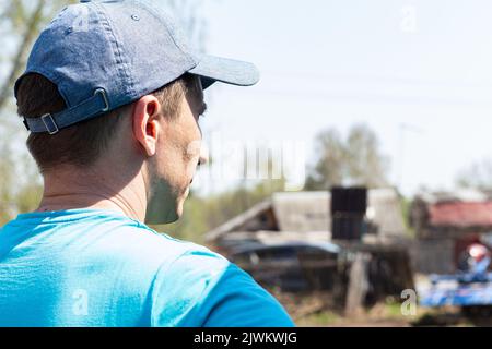 Nahaufnahme hübscher junger Bauer in blauem T-Shirt und Mütze steht auf dem Feld schaut auf sein Anwesen. Rückansicht. Selektiver Fokus, natürlicher Hintergrund auf s Stockfoto