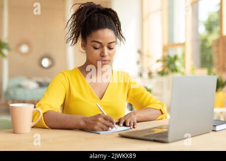 Fokussierte schwarze Studentin, die sich Notizen macht, Online-Vorträge auf dem Laptop ansieht, zu Hause online lernt, am Schreibtisch sitzt Stockfoto