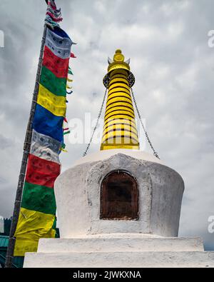 Farbenfrohe buddistische Stupa, flankiert von mehrfarbigen Gebetsfahnen, die an Holzmasten befestigt sind, alle unter wolkenbedecktem Himmel im Freien in Kalpa, Indien. Stockfoto