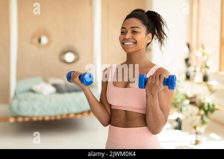 Porträt einer glücklichen afroamerikanischen Dame, die zu Hause ein Hanteltraining macht, an der Armstärke arbeitet und lächelt, Platz kopiert Stockfoto