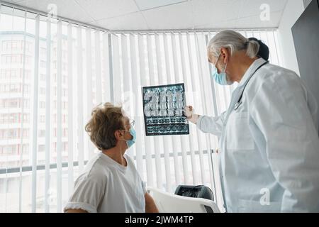 Arzt in Maske erklären kranken Patienten die Ergebnisse ihrer MRT- oder Röntgenaufnahme der Wirbelsäule Stockfoto