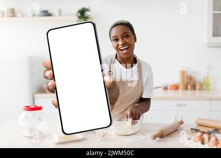 Kochen App. Fröhliche Schwarze Frau Backen In Der Küche Und Zeigt Unbeschriftete Smartphone Stockfoto