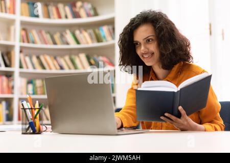 Fernschulungskonzept. Lächelnde, gemischte Rassendame, die in der Bibliothek sitzt und einen Laptop benutzt und Hausaufgaben für das College macht Stockfoto