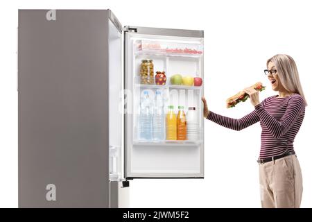 Frau, die ein Sandwich in der Hand hält und vor einem Kühlschrank steht, isoliert auf weißem Hintergrund Stockfoto