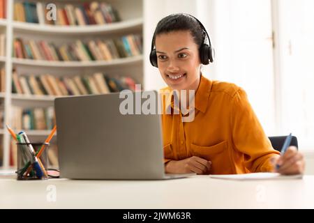 Glückliche Schülerin, die online mit einem Laptop lernt, fernab Unterricht hat und sich Notizen macht, in der Bibliothek sitzt Stockfoto