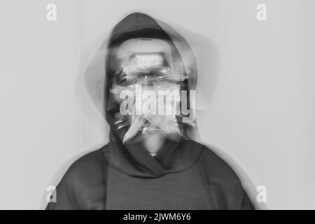 Ein Mädchen in einer Vollmaske aus Kunststoff und in einer Kapuze auf einem isolierten Hintergrund schüttelt ihren Kopf bei einer langen Exposition, Anonymität, Schreien und Verzweiflung, Psych Stockfoto