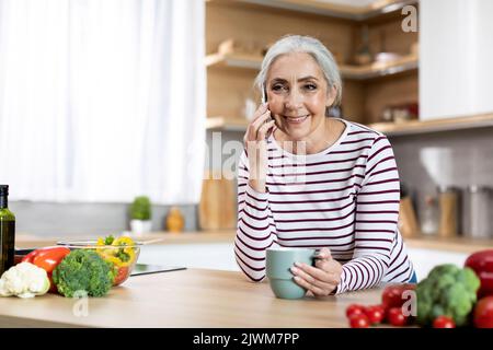 Lächelnde ältere Dame, die sich mit dem Handy unterhielt und Kaffee in der Küche trank Stockfoto