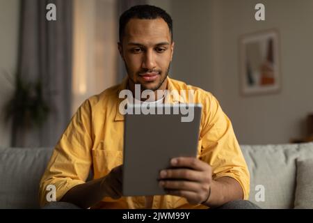 Fokussierter junger schwarzer Mann mit Tablet, der auf dem Sofa sitzt, online arbeitet oder studiert und zu Hause Geschäftstreffen abnimmt Stockfoto