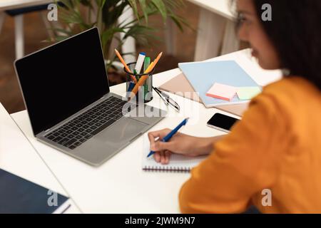 Online-Klassenkonzept. Schwarze Studentin, die im Notizbuch schreibt, Notizen macht und einen Laptop mit leerem Bildschirm für ein Modell verwendet Stockfoto
