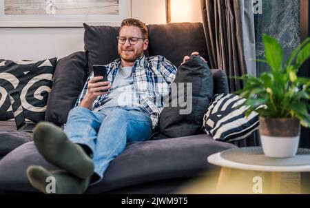 Entzückter erwachsener Mann, der auf der Couch lag und seine Freizeit mit seinem Mobiltelefon genoss. Stockfoto