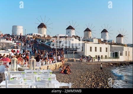 Zeitraffer, Touristen in der Nähe von Little Venice bewundern den Sonnenuntergang. Im Hintergrund die Windmühlen von Mykonos in Griechenland mit Panoramablick Stockfoto